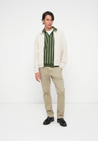 Młody mężczyzna stoi z rękami w kieszeniach, ubrany w beżowe spodnie, zieloną koszulę w paski, jasną kurtkę i czarne mokasyny, na tle białego tła.