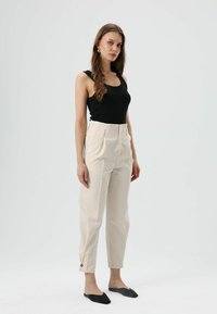 Schwarzes Tanktop, hoch taillierte cremefarbene Hose mit Falten, verkürzte Länge und Knopfdetail an der Manschette, kombiniert mit schwarzen spitzen Ballerinas.