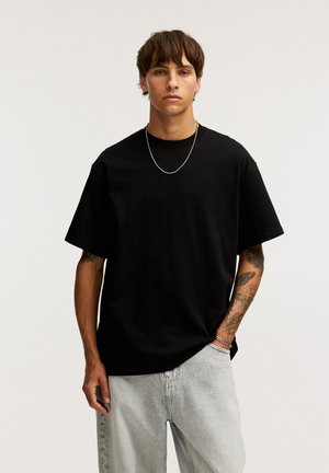 T-shirt en coton noir avec une coupe décontractée, des manches courtes et un col arrondi, associé à un jean en denim gris clair. Accentué par un simple collier en chaîne.