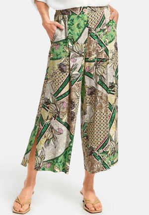 Pantalon à jambes larges et taille haute avec des motifs floraux et géométriques verts, beige et marron, fentes latérales et poches latérales, porté avec des sandales beiges.