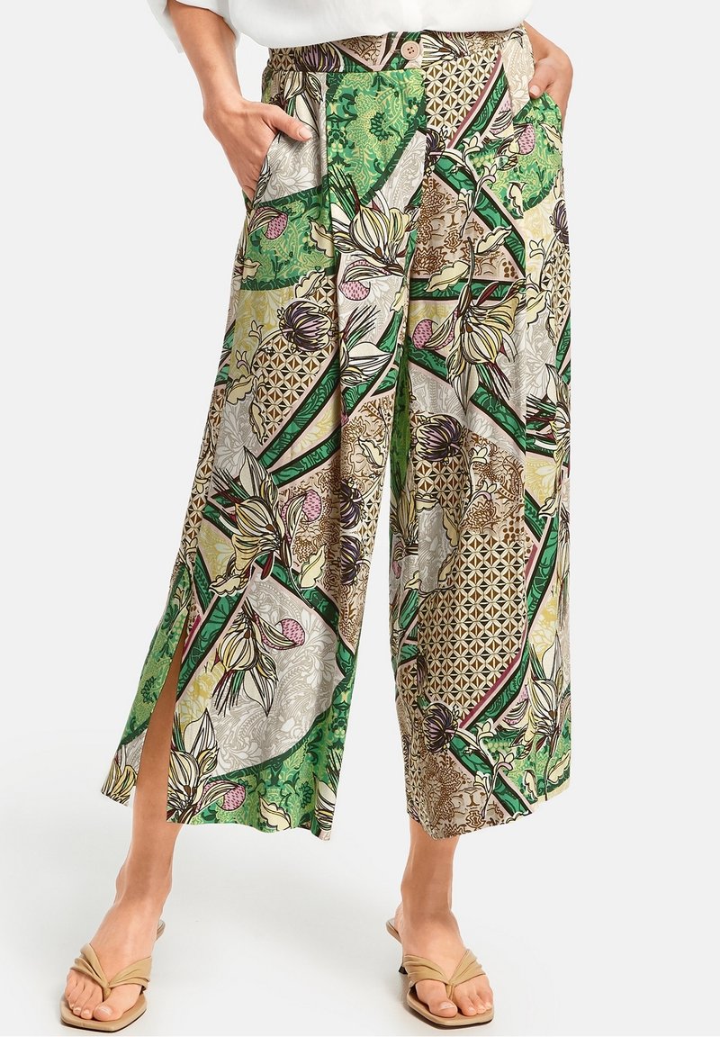 Pantalon à jambes larges et taille haute avec des motifs floraux et géométriques verts, beige et marron, fentes latérales et poches latérales, porté avec des sandales beiges.