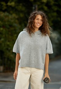 Világosszürke kötött pulóver rövid ujjakkal, csőnyakrész és bordázott szegéllyel, krémszínű nadrággal párosítva. Egyik kézben fekete csésze van.