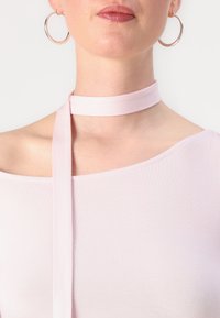 Gaiši rozā plecu bez piedurknēm tops ar garu auduma siksnu ap kaklu. Modelis valkā lielus sudraba riņķus ausīs. Gluda tekstūra.