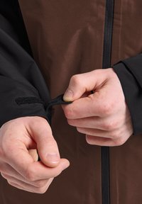 Giacca nera e marrone con zip frontale, polsini regolabili in Velcro e tessuto a trama liscia con finitura resistente all'acqua.