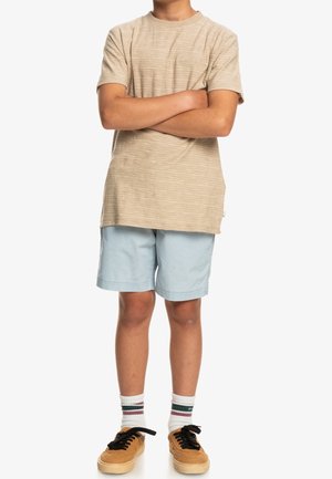 Barn med korslagte arme iført beige stribet t-shirt, lyseblå shorts, hvide sokker med striber og brune sneakers.