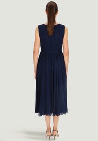 Granatowa plisowana sukienka z dopasowanym stanem i spódnicą do kolan. Przezroczysta tekstura, okrągły dekolt i gładkie wykończenie. Nosi ją w połączeniu z sandałami na obcasie.