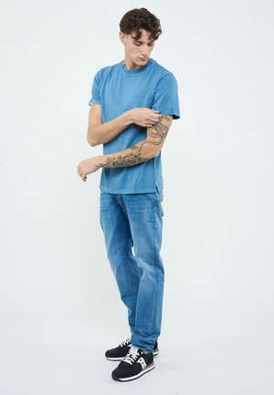 Giovane uomo con maglietta blu e jeans che mostra dettagliati tatuaggi di serpente e fiori sul braccio sinistro su uno sfondo semplice.