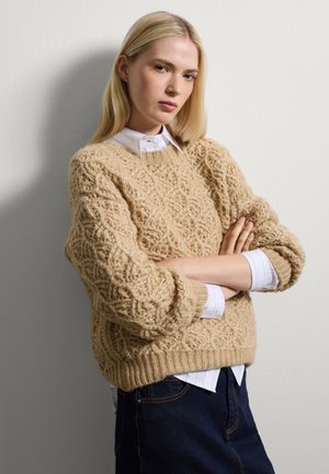 Maglione beige lavorato a maglia con motivo testurizzato, scollo rotondo e orlo a coste. Indossato sopra una camicia bianca con colletto, abbinato a jeans scuri.