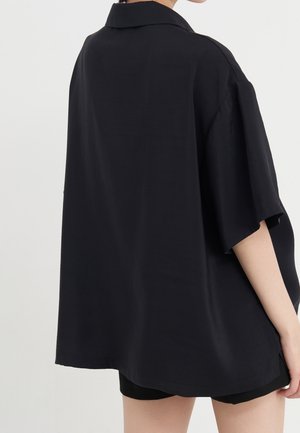 Mujer con camisa negra de manga corta y cuello, de corte holgado, vista desde atrás, combinada con pantalones cortos negros sobre un fondo liso.