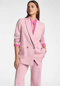 Blazer rosa doppiopetto con bottoni dorati, caratterizzato da una vestibilità sartoriale e maniche arrotolate, abbinato a una camicia rosa con colletto sotto.
