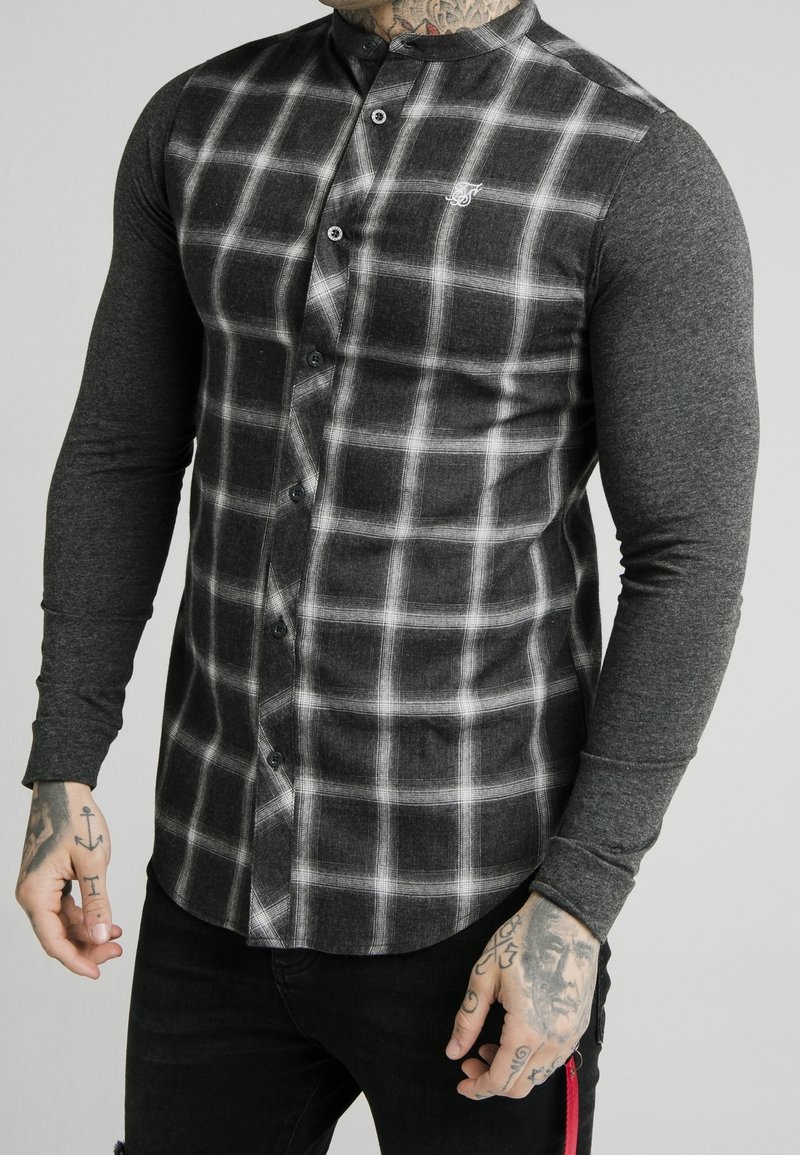 Chemise à carreaux gris avec col mandarin, manches longues gris foncé contrastantes et boutons noirs visibles.