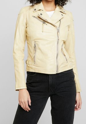 Vrouw draagt een lichtbeige leren bikerjack met asymmetrische rits en zwarte spijkerbroek tegen een effen achtergrond.