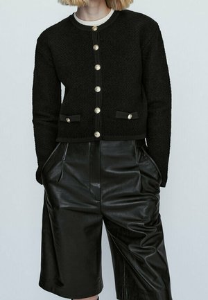 Woman wearing a black textured buttoned cardigan with gold buttons, paired with black high-waisted leather shorts with side pockets.