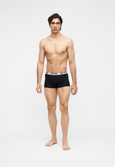 Νέος άνδρας που στέκεται ξυπόλητος φορώντας μαύρα boxer Moschino με λευκή ζώνη στηριγμένη σε μονόχρωμο φόντο.