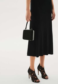 Fekete magas sarkú szandál bonyolult kivágott virágmintával, egy apró fekete bársony kézitáskával párosítva, amely ezüst színű kiegészítőkkel és fogantyúval rendelkezik.