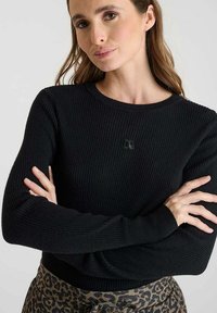 Pull noir côtelé à manches longues et col rond, présentant un logo en relief subtil sur la poitrine. Stylisé sur un pantalon imprimé.