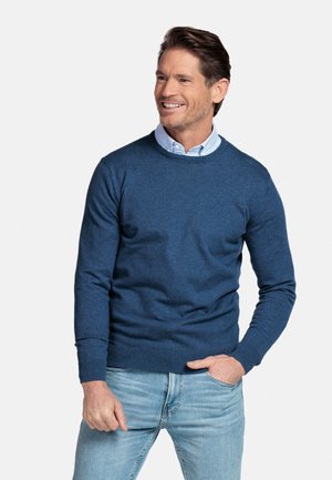 Man die lacht, draagt een blauwe trui met ronde hals over een lichtblauw overhemd en lichtgewassen spijkerbroek, staand tegen een effen witte achtergrond.