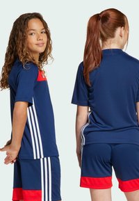 Modrý sportovní komplet skládající se z trička s krátkým rukávem, červenými akcenty a bílými pruhy, doplněný odpovídajícími šortkami. Hladká textura látky.