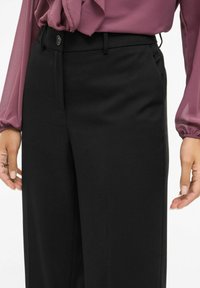 Črne hlače s prilagojeno prileganjem, z gumbom za zapiranje, ravnim sprednjim delom in žepi, kombinirane s vijolično, rahlo prozorno bluzo.