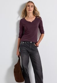 Langdurige bordeaux top met een henley-halslijn en drie knopen, gecombineerd met donkergrijze jeans en een lichtbruine suède schoudertas.