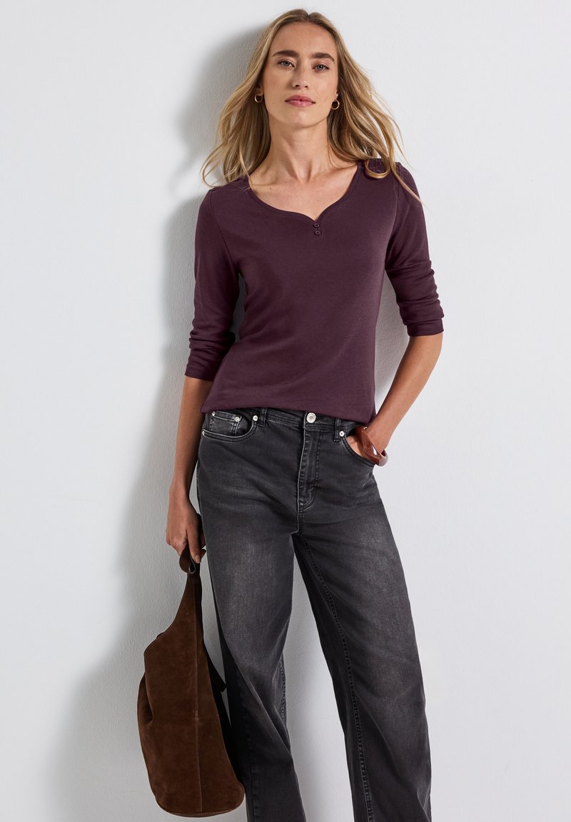 Langdurige bordeaux top met een henley-halslijn en drie knopen, gecombineerd met donkergrijze jeans en een lichtbruine suède schoudertas.