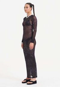 Schwarzes, transparentes Kleid mit langen Ärmeln, floralem Muster und textuellen Elementen. Figurbetontes Design, kombiniert mit schwarzen Plateausandalen. Glatte Textur.