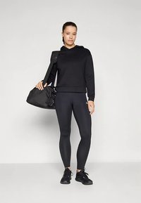 Zwarte hoodie met een cropped design, gecombineerd met zwarte leggings. Het model draagt een zwarte duffeltas en zwarte sneakers.