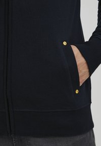 Felpa con zip di colore navy, con polsini e orlo ribattuti, presenta una tasca con dettagli di bottoni gialli. Il materiale sembra morbido e strutturato.