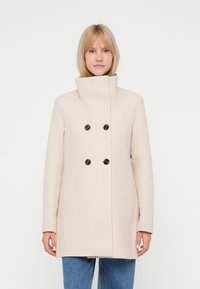 Cappotto in misto lana beige con colletto alto, chiusura doppiopetto con bottoni neri e tasche laterali, abbinato a pantaloni in denim blu.