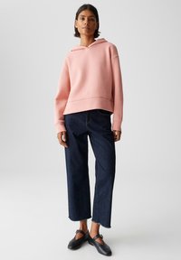 Cropped hoodie rosa chiaro con cordoncino, abbinato a jeans larghi blu scuro e scarpe slip-on nere, su uno sfondo neutro.