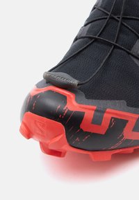 Schwarzer Sportschuh mit strukturiertem Obermaterial, roten Akzenten und gemustertem Sohlenprofil. Enthält Logo und elastische Schnürsenkel zur Anpassung der Passform.