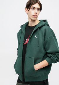 Felpa con zip di colore verde scuro in tessuto morbido, con tasca a marsupio, caratterizzata da un logo bianco di Jordan sulla parte superiore sinistra. T-shirt nera sotto.