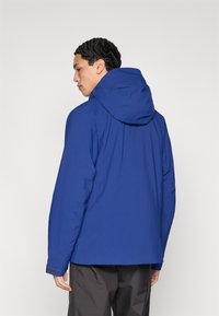 Blå huvad jacka med slät textur, utrustad med elastiska muddar och en avslappnad passform. Bakifrån visas en minimalistisk design utan synliga logotyper.