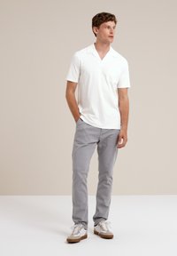 Camisa polo blanca de manga corta con bolsillo, combinada con pantalones gris claro y zapatillas blancas con detalles en marrón, sobre un fondo neutro.