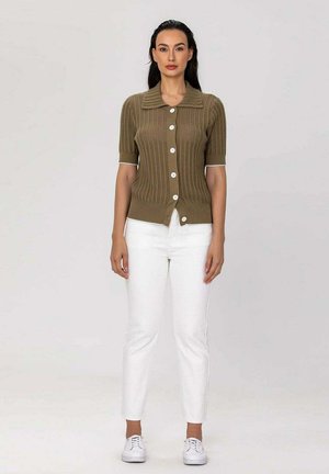Femme debout, portant un pull en maille marron à manches courtes boutonné, pantalon blanc taille haute et baskets blanches, fond neutre en studio.