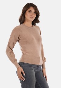 Maglione beige lavorato a maglia con texture a coste, scollo rotondo e maniche aderenti. Indossato con jeans grigio scuro per un contrasto.