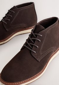 Braune Chukka-Stiefel aus Wildleder mit grauen Schnürsenkeln, einer beigen Zwischensohle und kontrastierenden Steppnähten. Das Design umfasst eine abgerundete Spitze und einen knöchelhohen Schaft.