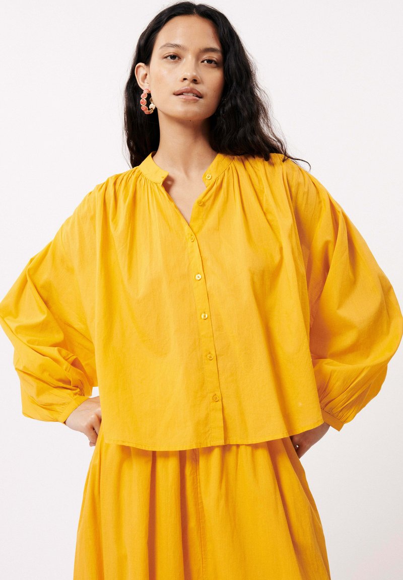 Chemisier jaune à boutons avec une coupe décontractée, des manches bouffantes et un col froncé. Tissu texturé avec un drapé doux, associé à une jupe fluide.