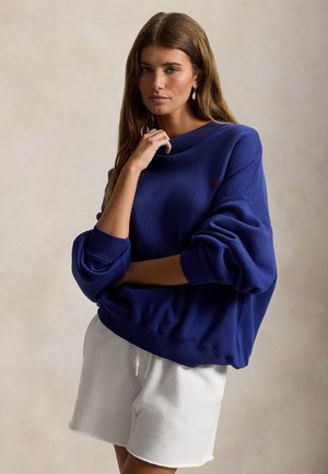 Laivaston sininen ylisuurikokoinen collegepaita, jossa on pyöreä pääntie ja laskostetut olkapäät, resorit hihansuissa sekä pieni logoyksityiskohta. Yhdistetty valkoisten shortsien kanssa.