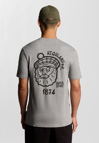 Grijze T-shirt met korte mouwen met een zwart cartoonafbeelding van een baardige figuur die een geruite hoed draagt, met de tekst "HIGHLANDER" en "1874".