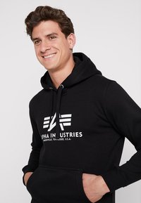 Zwarte hoodie met een voorkant kangaroo zak, witte logo tekst "ALPHA INDUSTRIES" en ontwerpaccenten. Zachte stof met trekkoorden voor een verstelbare pasvorm.
