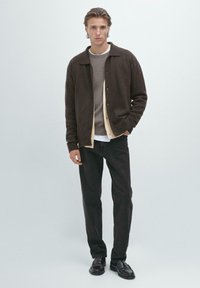 Mladý muž stojí, oblečený v vrstvené hnědé bundě, béžovém svetru, šedém svetru, černých kalhotách a černých kožených botách na jednobarevném světlém pozadí.