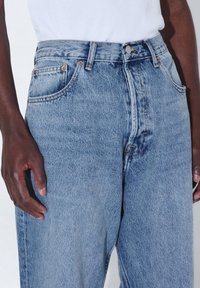 Jeans de mezclilla azul claro con cintura alta, cinco bolsillos y herrajes de cobre. Tejido texturizado con un corte clásico y detalles de desgaste sutil.