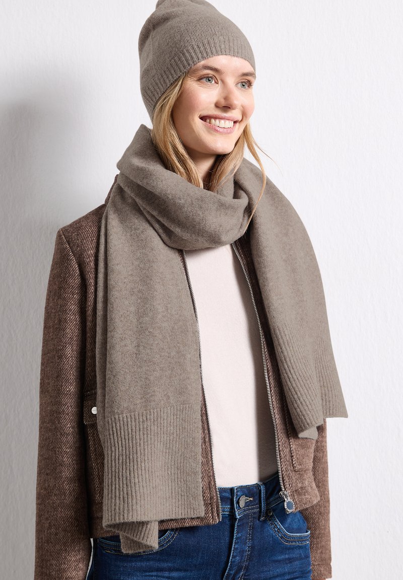Femme souriante portant un bonnet en tricot taupe et une écharpe assortie, une veste marron texturée, un haut blanc et un jean bleu devant un mur blanc.