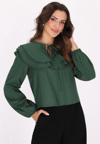 Grüne Bluse mit Rüschendetails und Pomponbesatz, mit einem Schlüssel-ausschnitt und langen Ärmeln, getragen mit schwarzen Hosen.