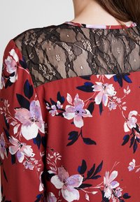 Röd blus med blommönster och en strukturerad svart spetsdetalj, med rosa och vita blommor, marinblå blad och en slät tygfinish.