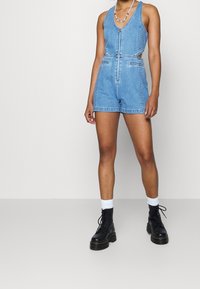 Jeansöveralls i ljusblått med dragkedja fram, urtagna sidor och två framfickor. Bärs med svarta snörkängor och vita strumpor.