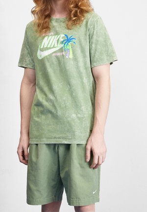 Groene tie-dye T-shirt met een levendig Nike-logo en palmboomgraphic, gecombineerd met bijpassende groene shorts met een kleine Nike swoosh.