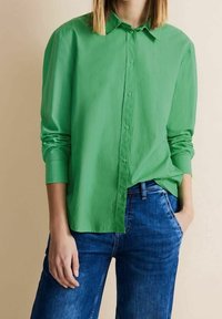 Grön skjorta med knappar, tillverkad av lätt tyg, med krage och lång ärm, ihop med blå denimjeans.