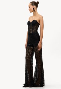 Femme portant un corset bustier en dentelle noire transparente et un pantalon large assorti, stylisée avec de longues boucles d'oreilles à pampilles noires et des chaussures noires.
